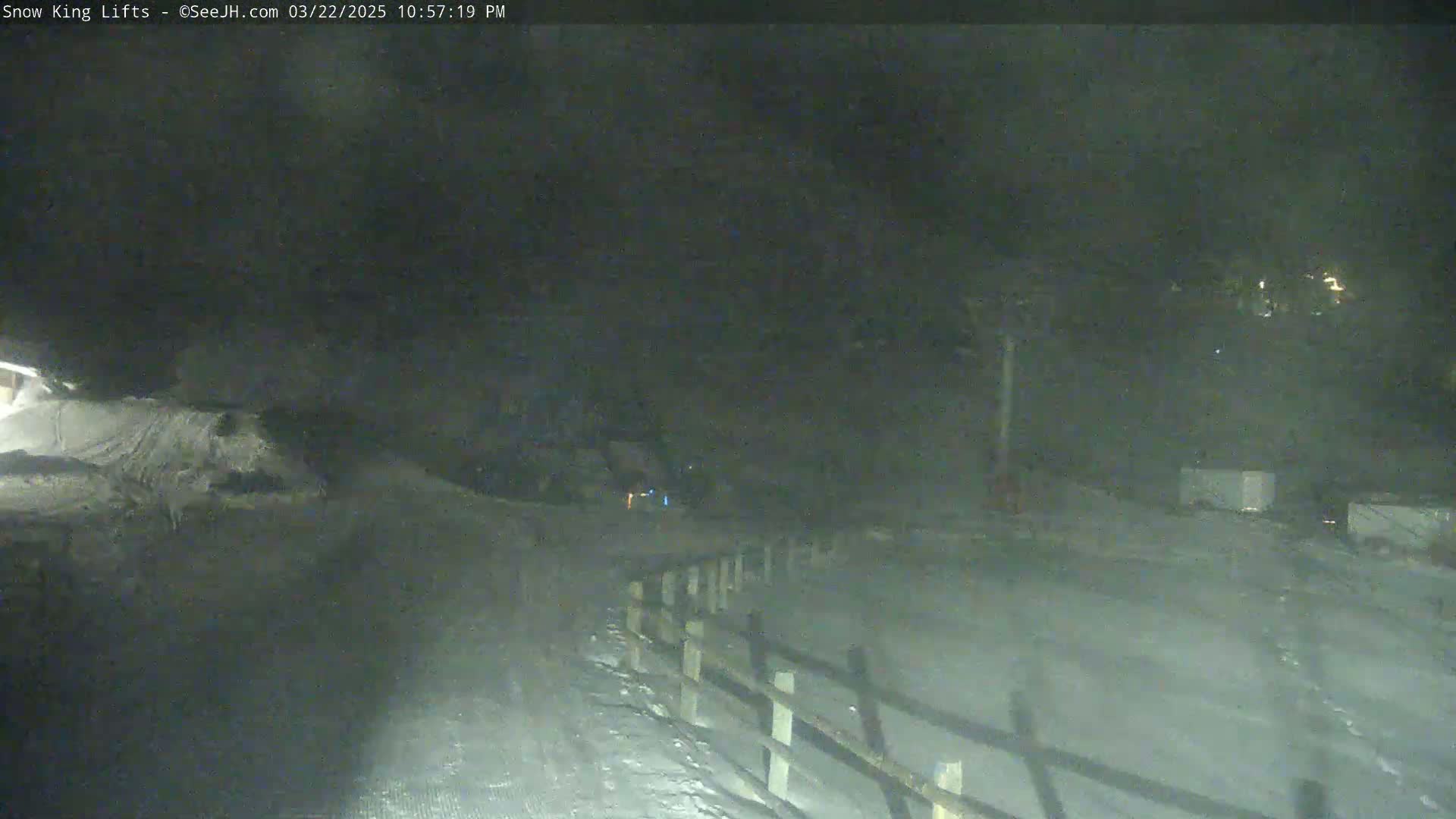 A snow-covered area at night is dimly lit, showing a wooden fence and a few indistinct objects in the distance.