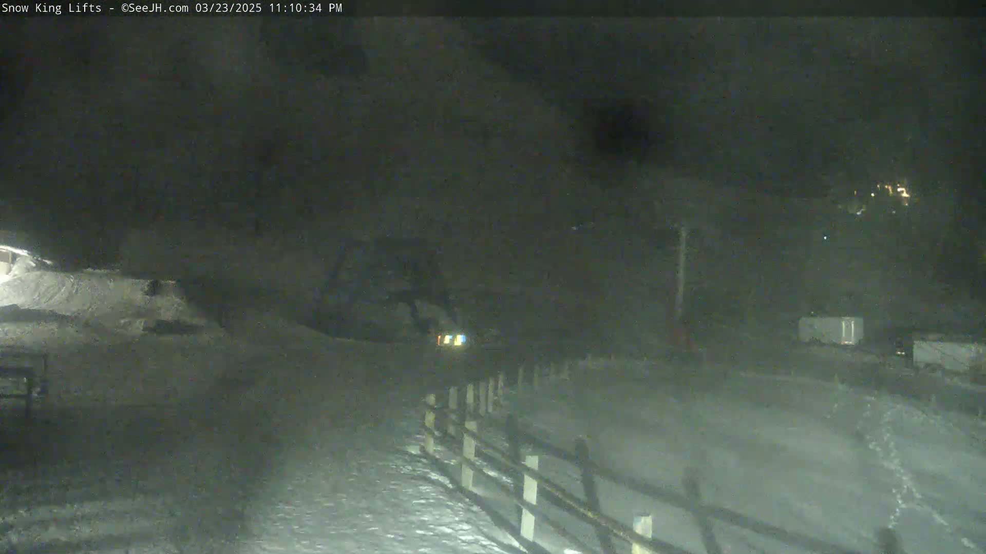 A nighttime snowy scene shows a snow-covered area with a fence, a vehicle with headlights on, and some distant lights.