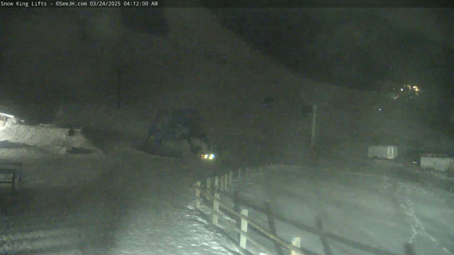A nighttime, snow-covered scene shows a vehicle's headlights in the distance, a snow-covered area in the foreground, and a wooden fence along the right.