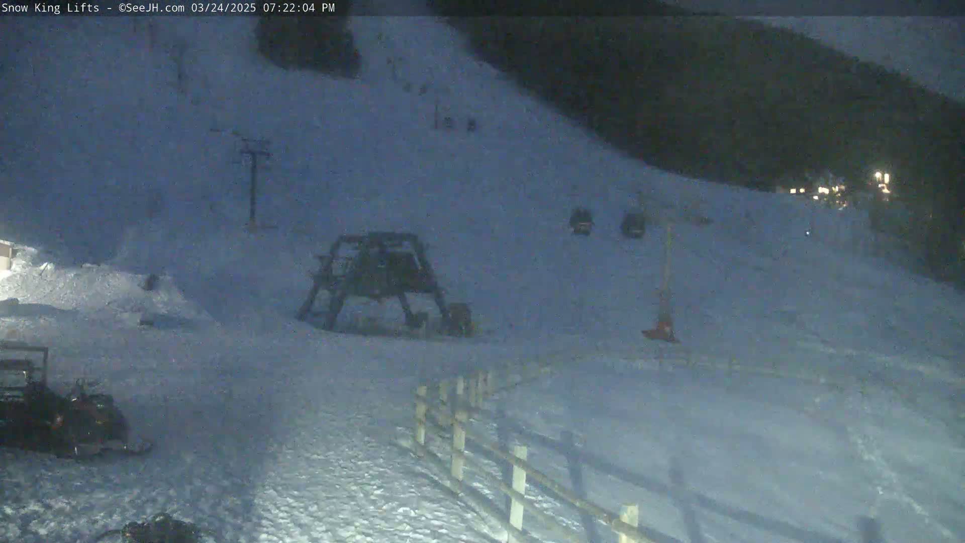 A snow-covered ski slope at night, with a ski lift and several people visible in the distance.