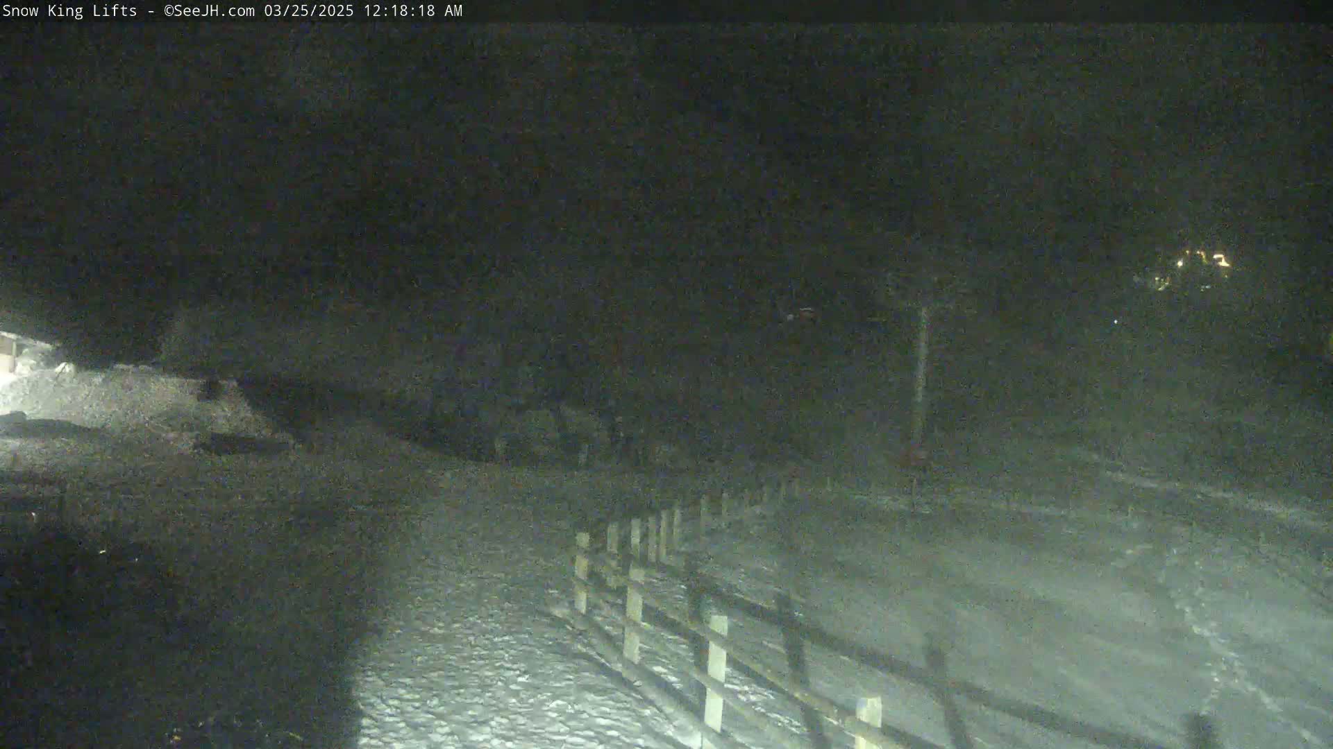 A nighttime, snowy scene shows a wooden fence alongside a snow-covered path leading towards some distant lights.