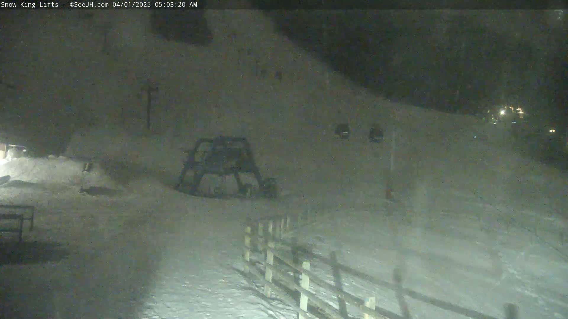 A nighttime, snowy scene shows a snowcat parked near a wooden fence, with a gently sloping snow-covered hill extending into the background.