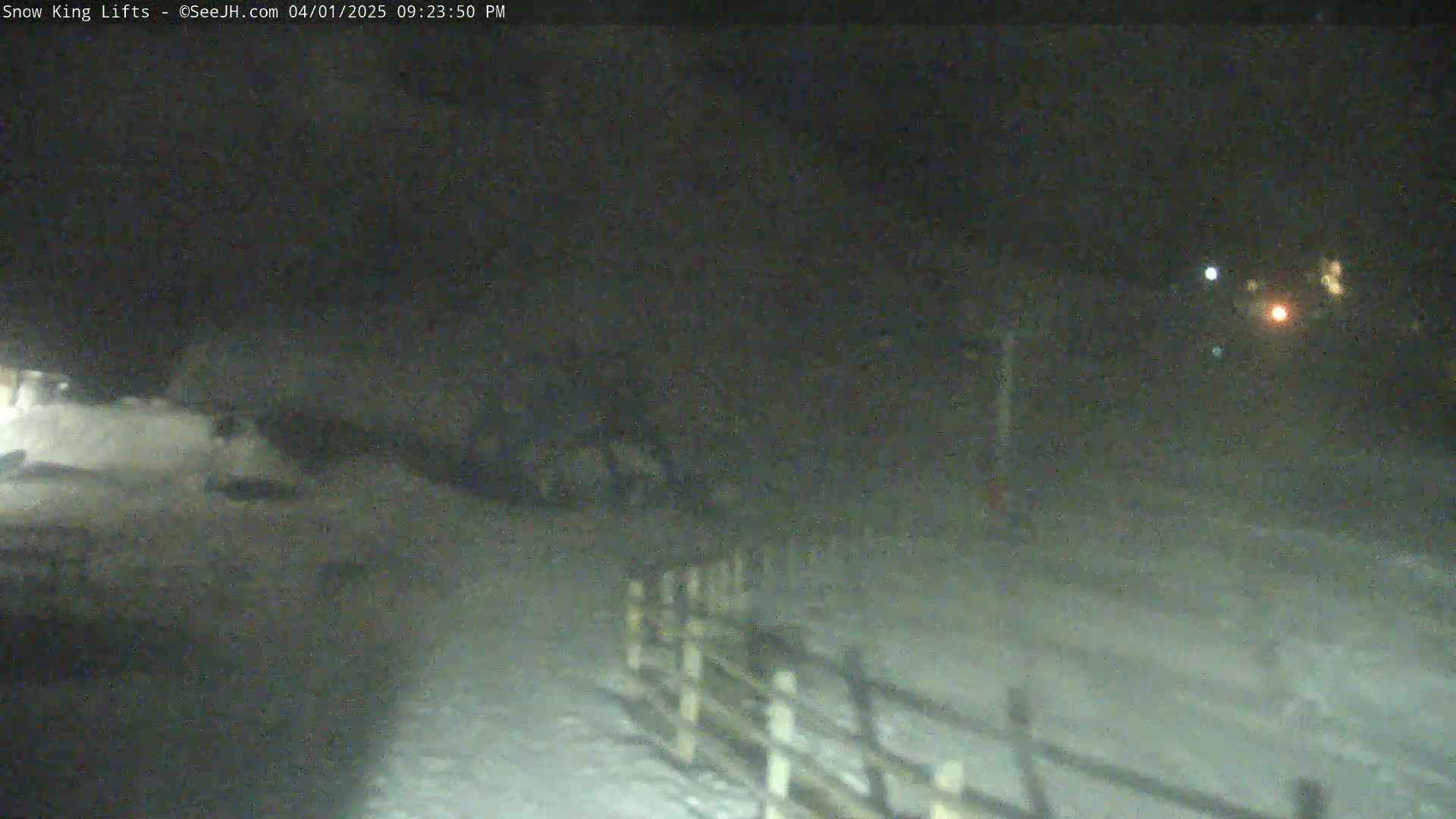 A snow-covered area at night, illuminated by distant lights, with a wooden fence visible in the foreground.