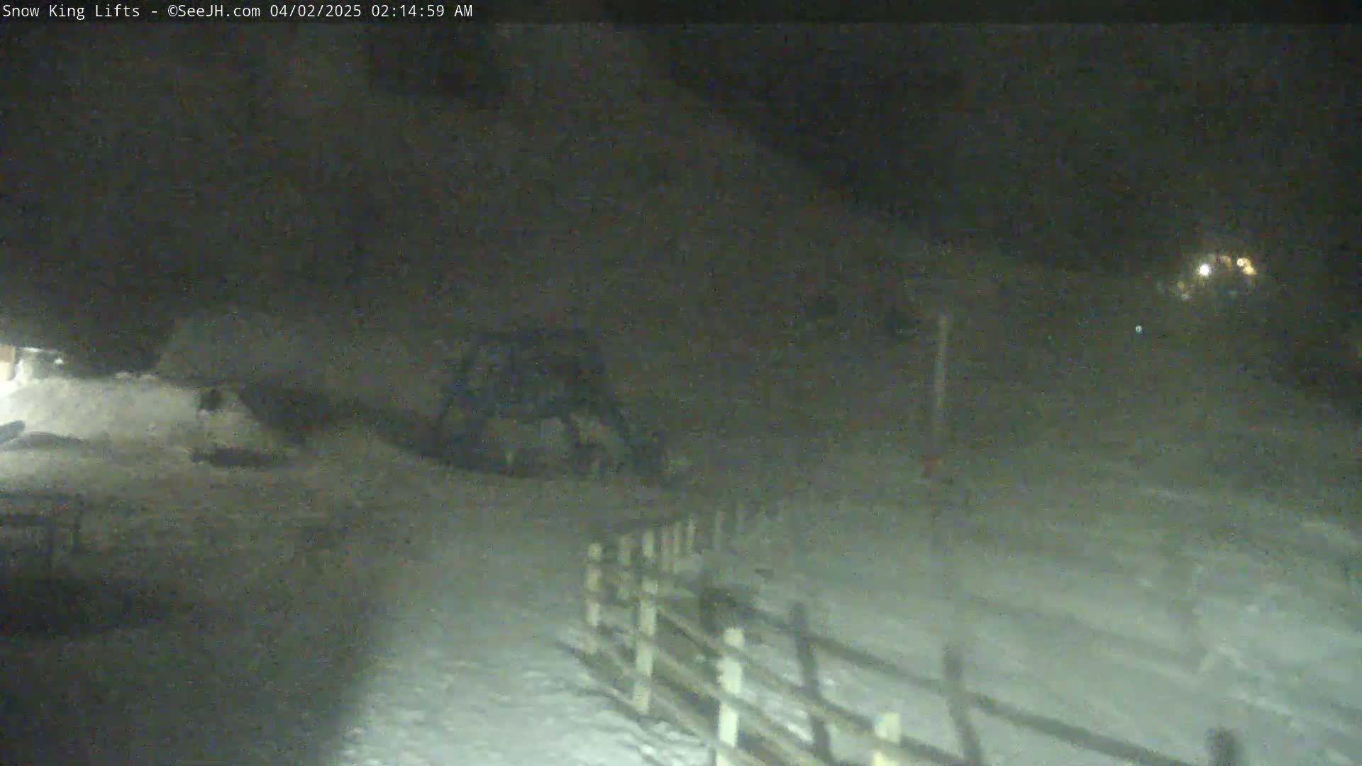 A dark, snowy scene at night shows a snow-covered ground with a wooden fence and some indistinct objects in the distance under the cover of darkness.