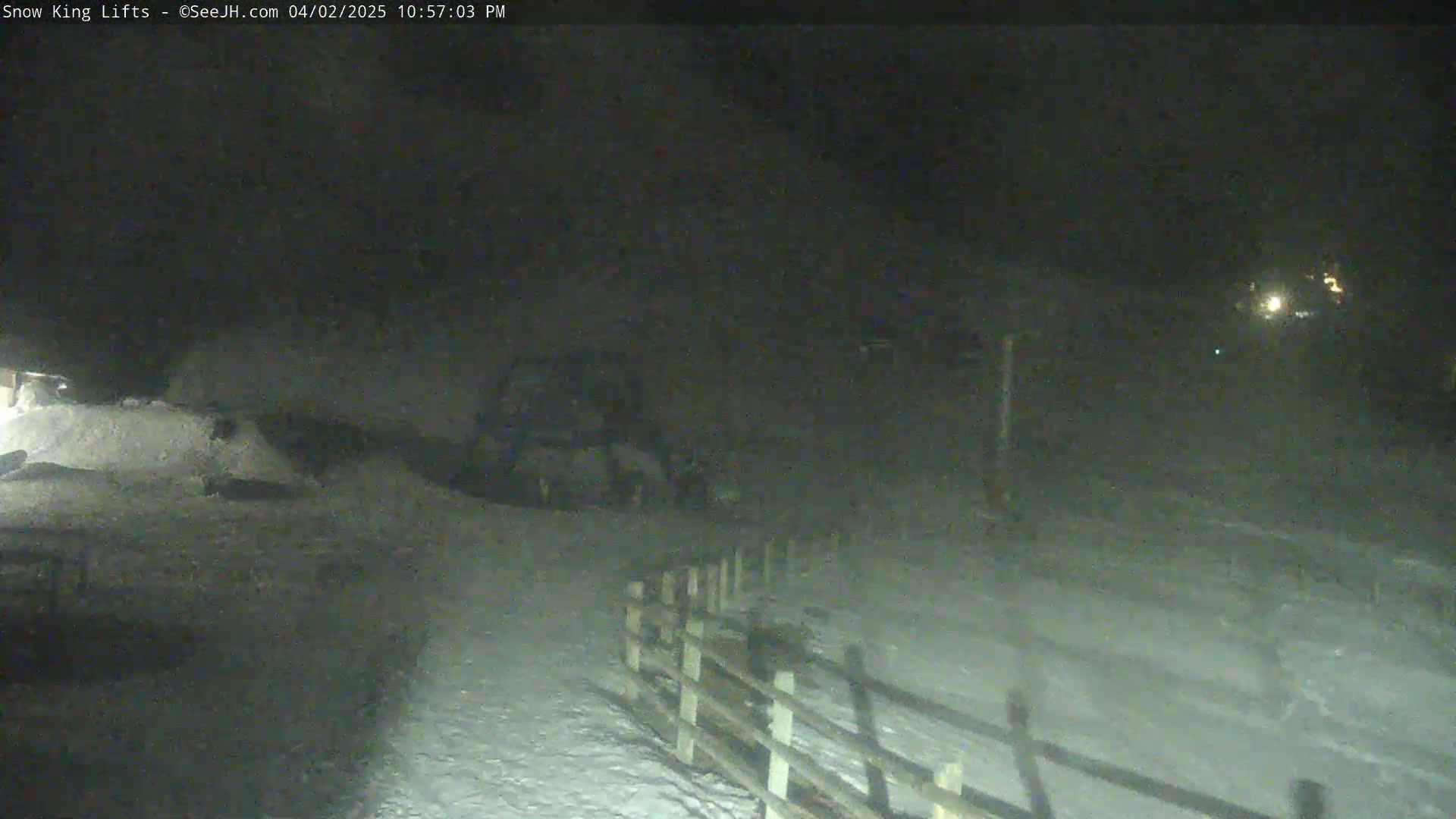 A snow-covered area at night is illuminated by a distant light source, showing a piece of equipment and a wooden fence.
