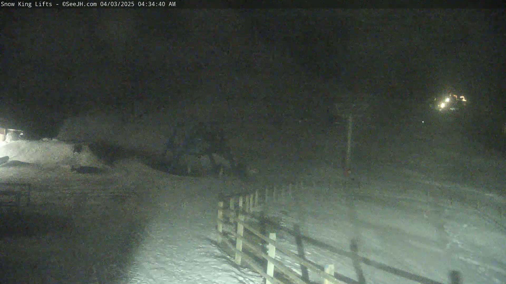 A snow-covered area at night is shown, with a wooden fence and distant lights visible in the darkness.