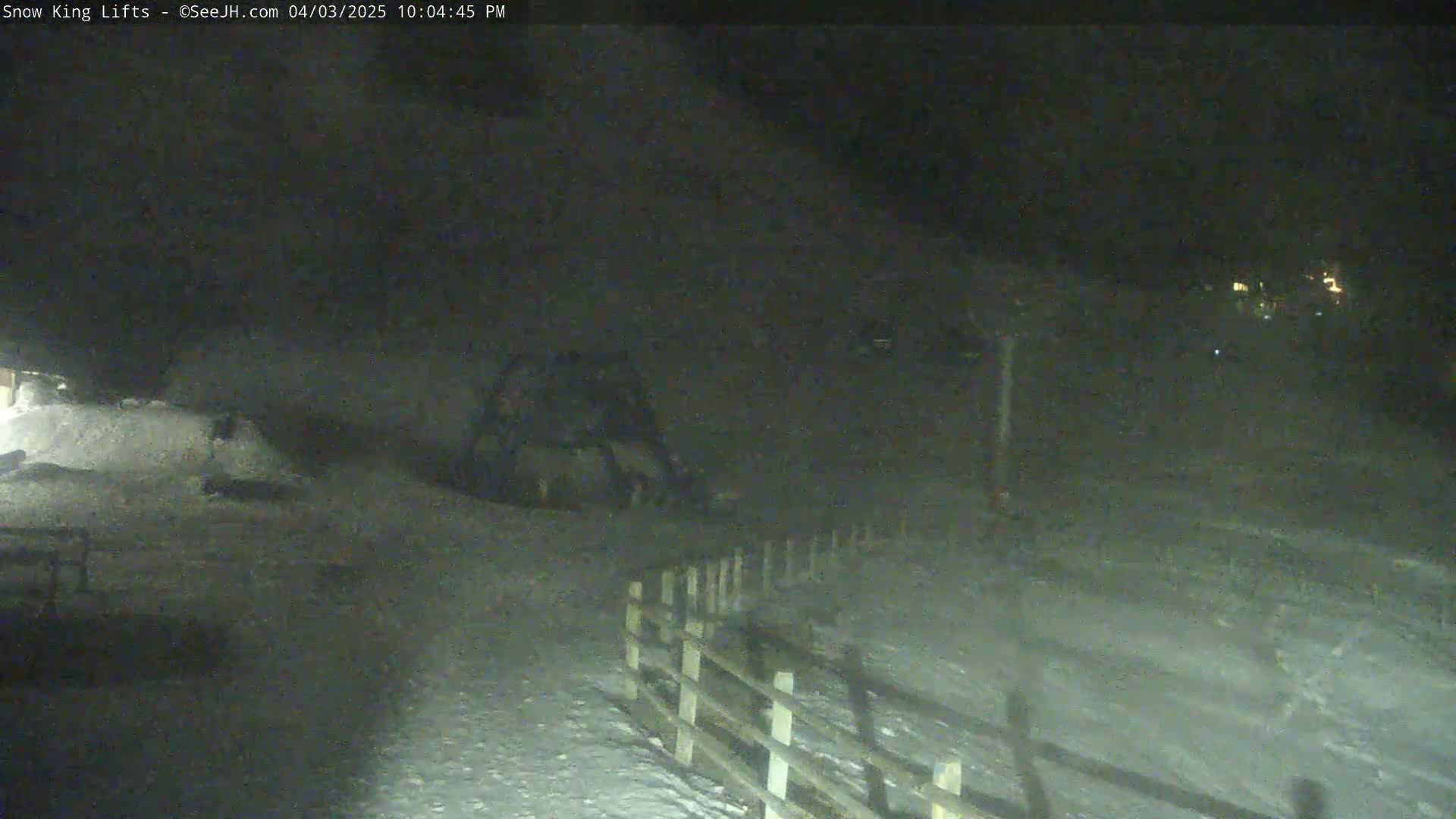 A snow-covered landscape is visible at night, with a wooden fence in the foreground and faint lights in the distance.