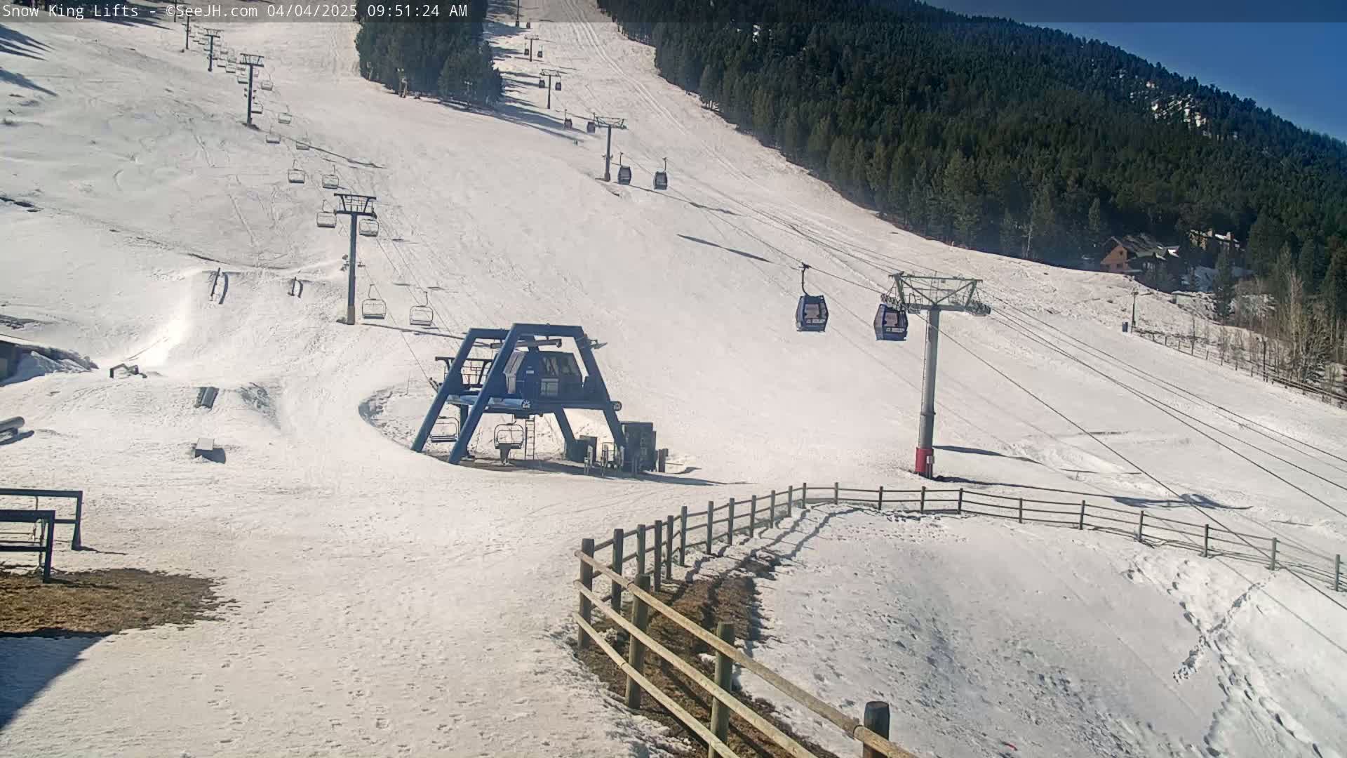 A snow-covered ski slope with several chairlifts and a gondola, under sunny skies, is bordered by a wooden fence and a coniferous forest.