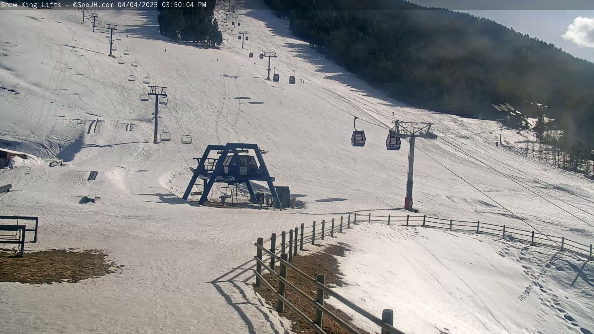 A snow-covered ski slope with several chairlifts, a small building, and a wooden fence under a sunny sky.