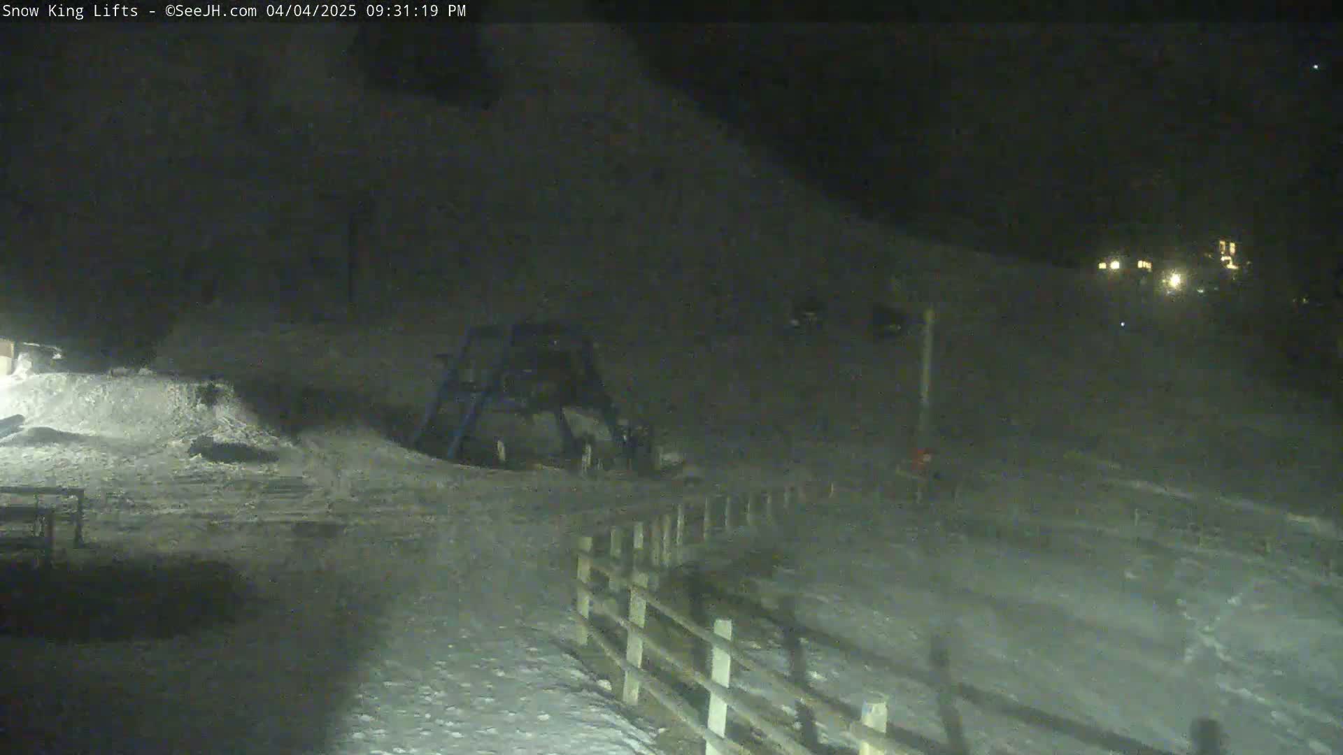 A snow-covered area at night, illuminated by faint lights in the distance, shows a wooden fence and what appears to be playground equipment.