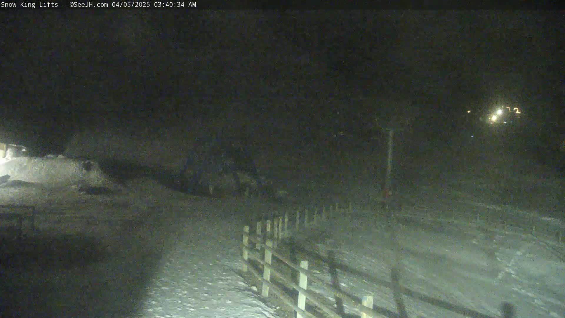 A snow-covered area at night, illuminated by distant lights, features a wooden fence and appears to be a lightly trafficked outdoor location.