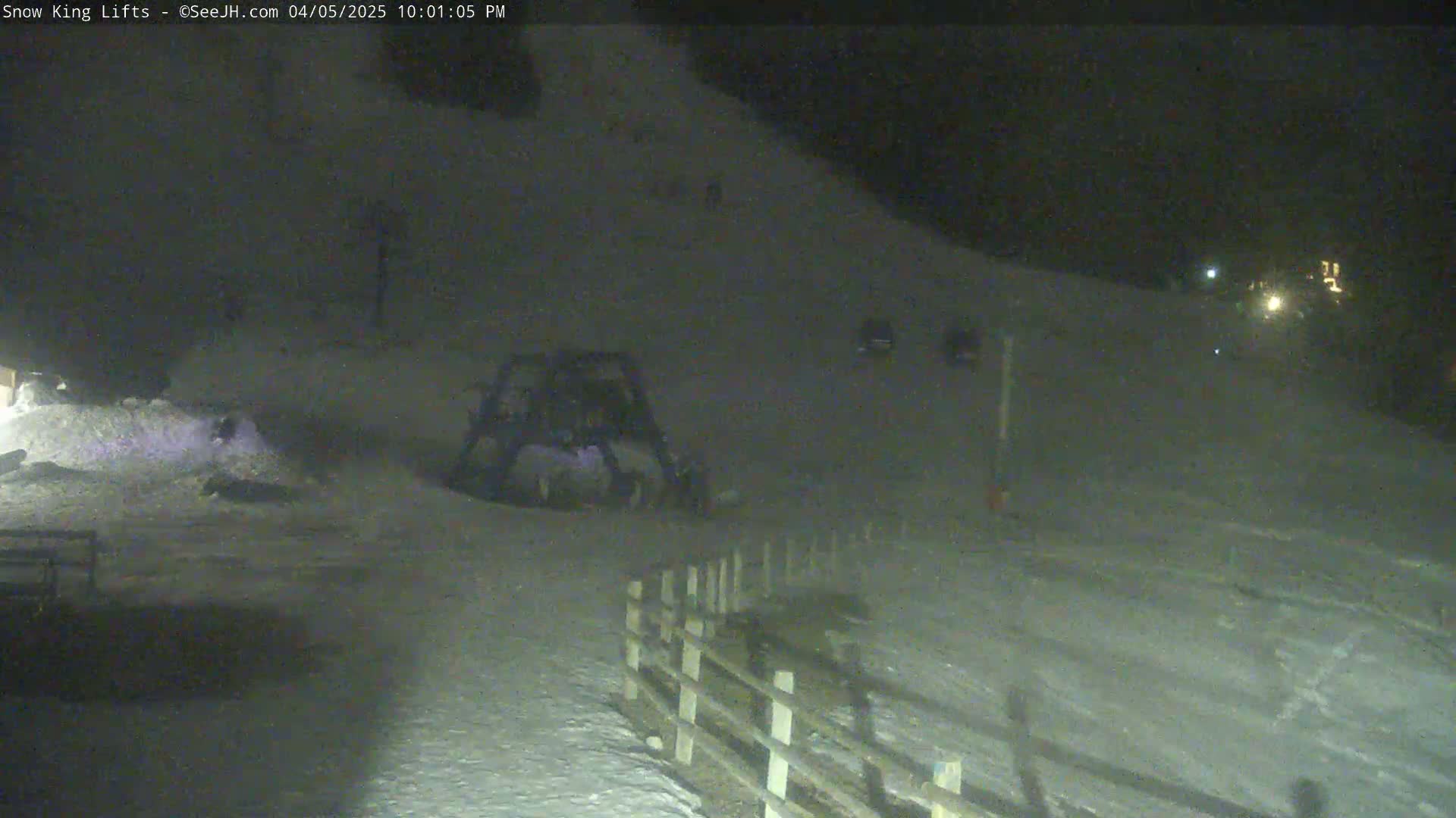 A snow-covered slope at night is illuminated by a few distant lights, with a piece of snow-grooming equipment parked near a wooden fence.