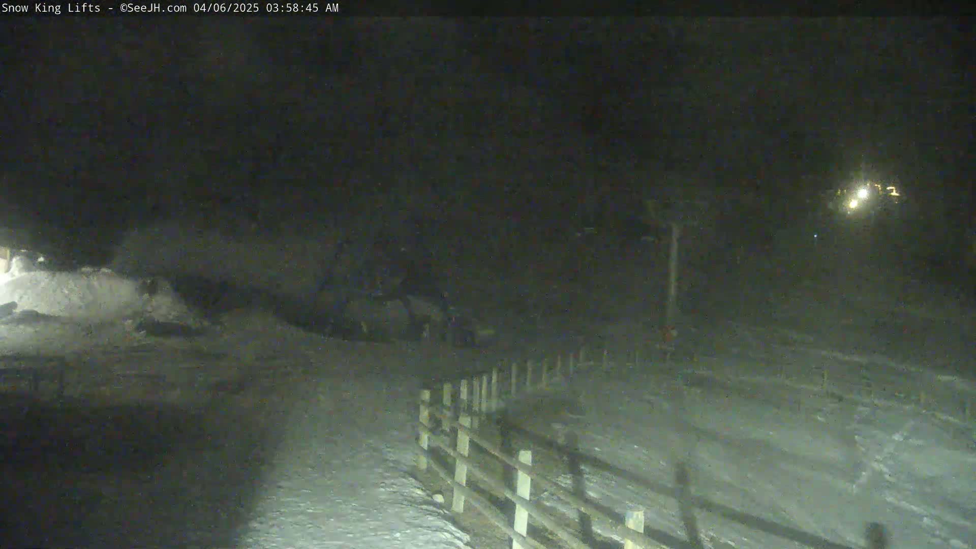 A snow-covered area at night, illuminated by distant lights, with a wooden fence in the foreground.