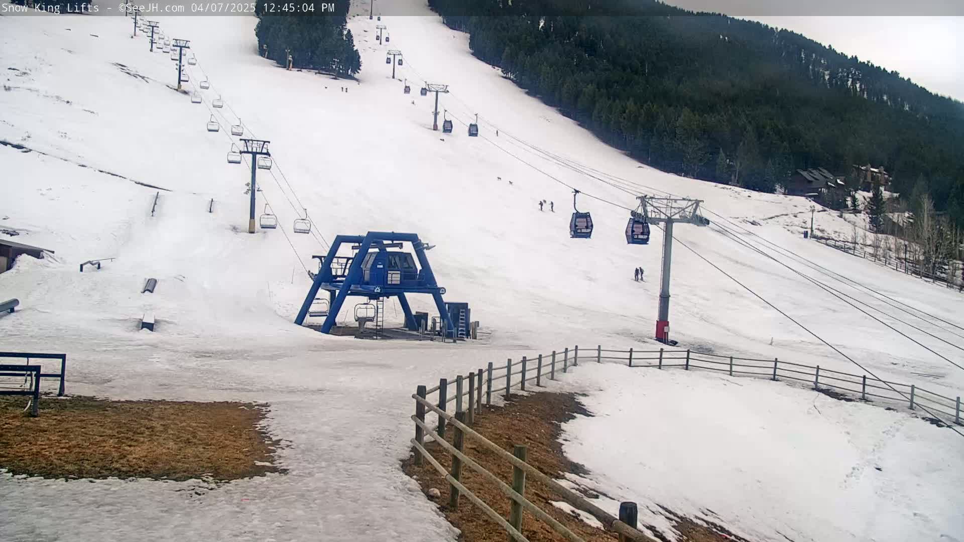 A ski lift with several chairlifts ascending a snow-covered slope, backed by a dark evergreen forest under an overcast sky.