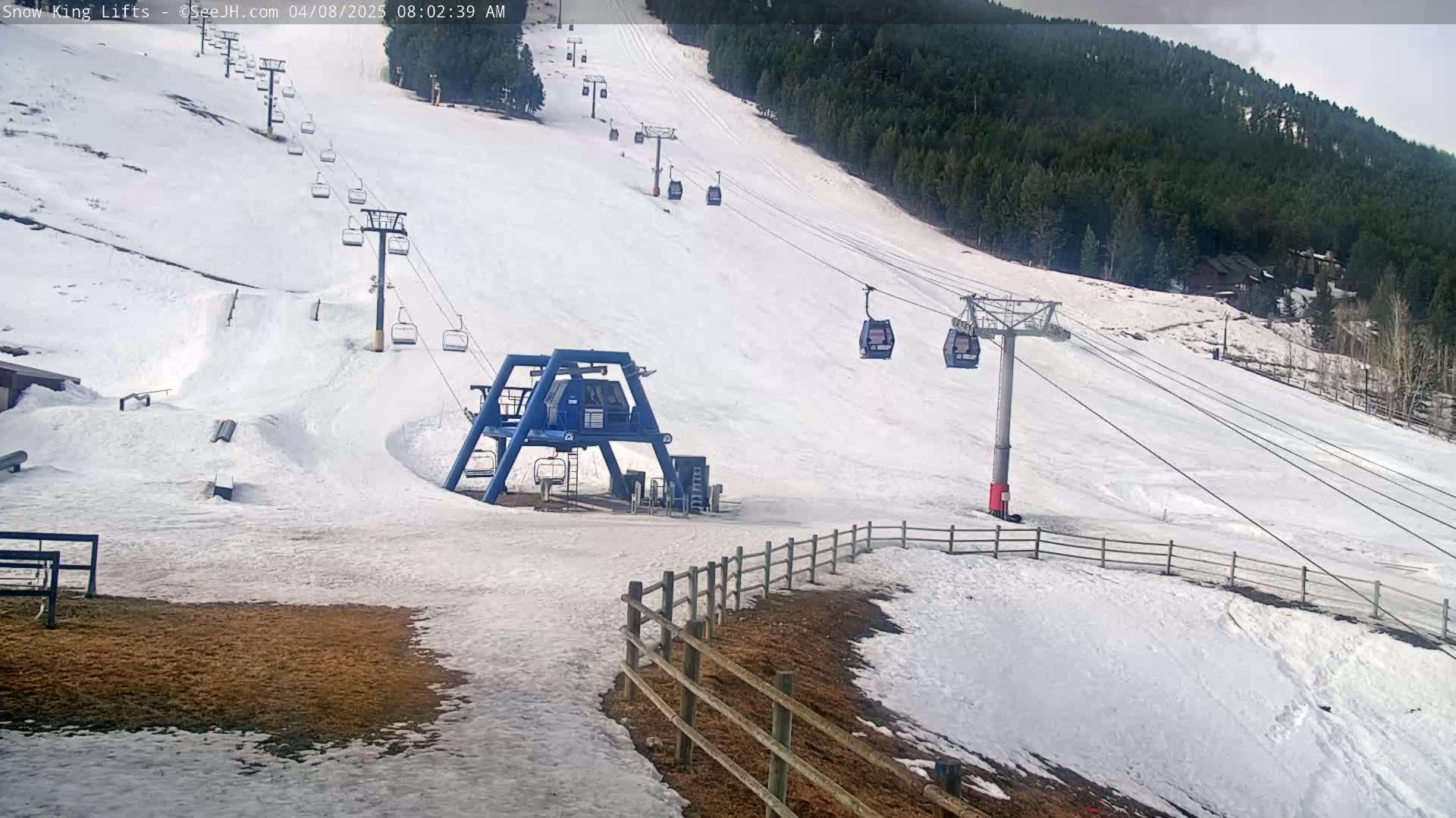 A ski lift base station and several chairlifts ascend a snow-covered ski slope, backed by a coniferous forest under a mostly cloudy sky.