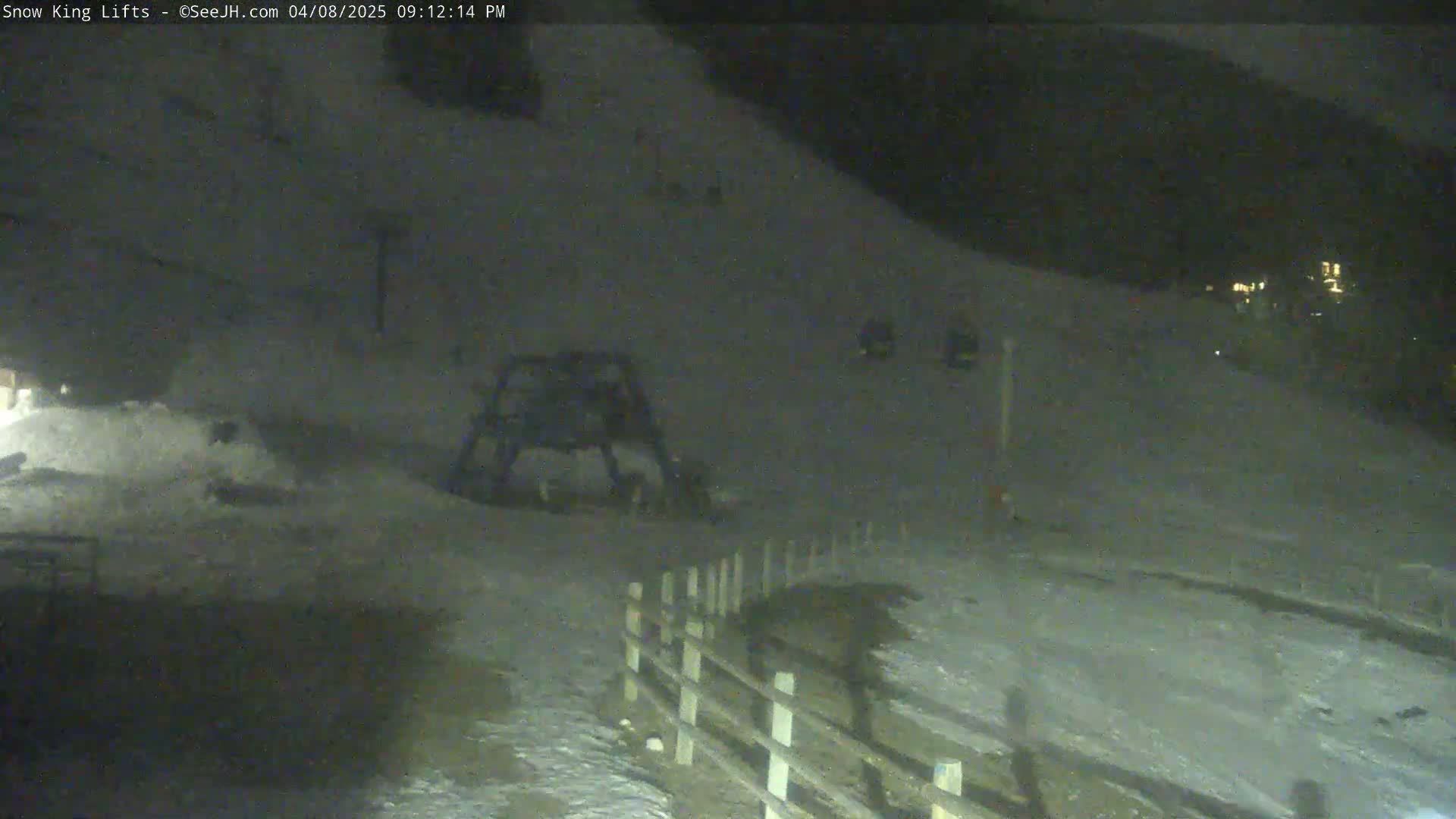 A nighttime, snow-covered slope with a ski lift and a wooden fence is visible under dark conditions.