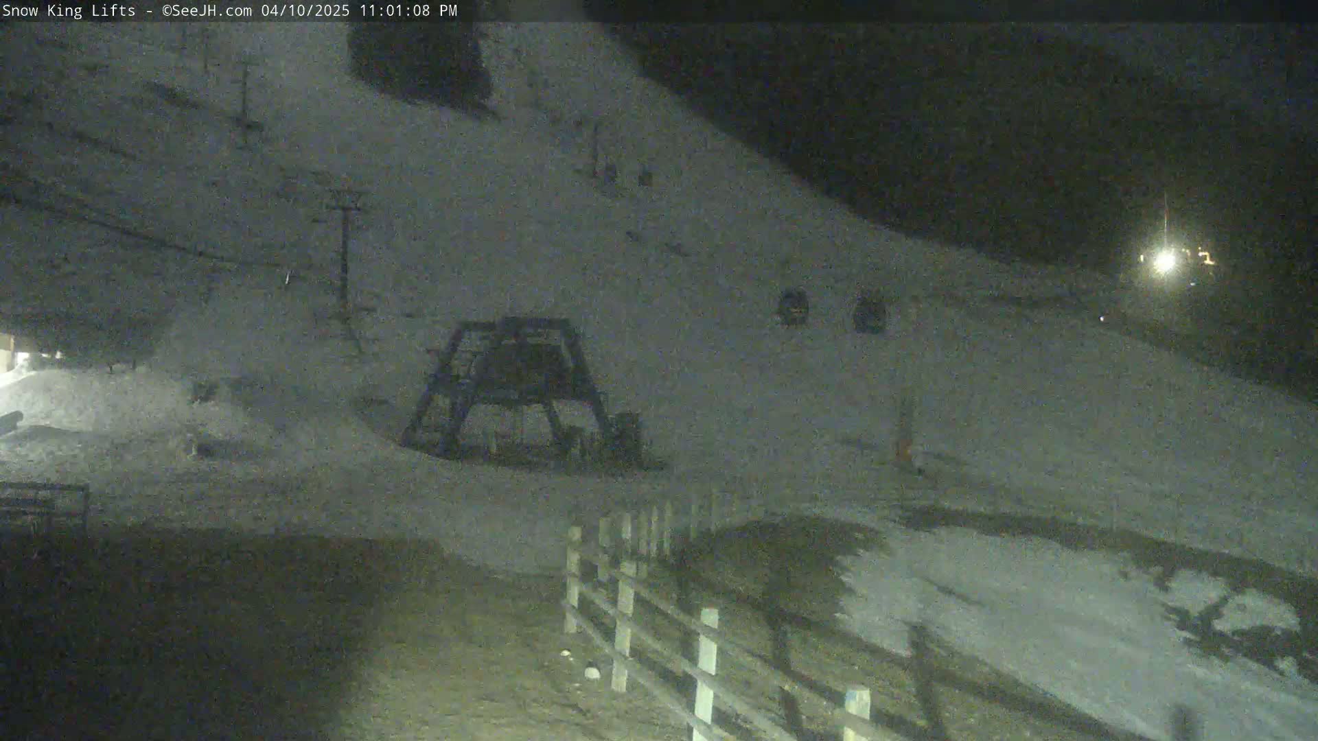 A nighttime view of a snow-covered ski slope with a ski lift and a fence at the bottom, illuminated by a light source in the distance.