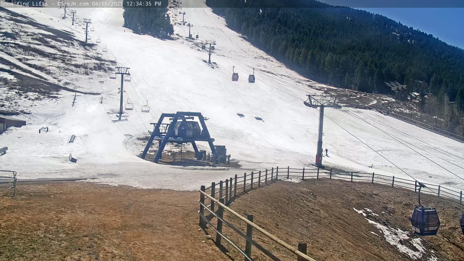 A ski lift with several empty chairs and gondolas ascends a snow-covered slope on a sunny day, with a treeline visible in the background.