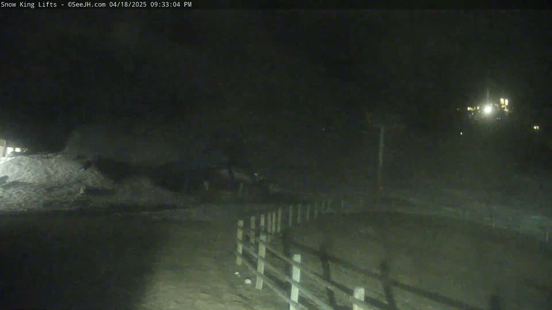 A nighttime, low-light view shows a snow-covered area near a wooden fence, with dimly lit structures visible in the distance.