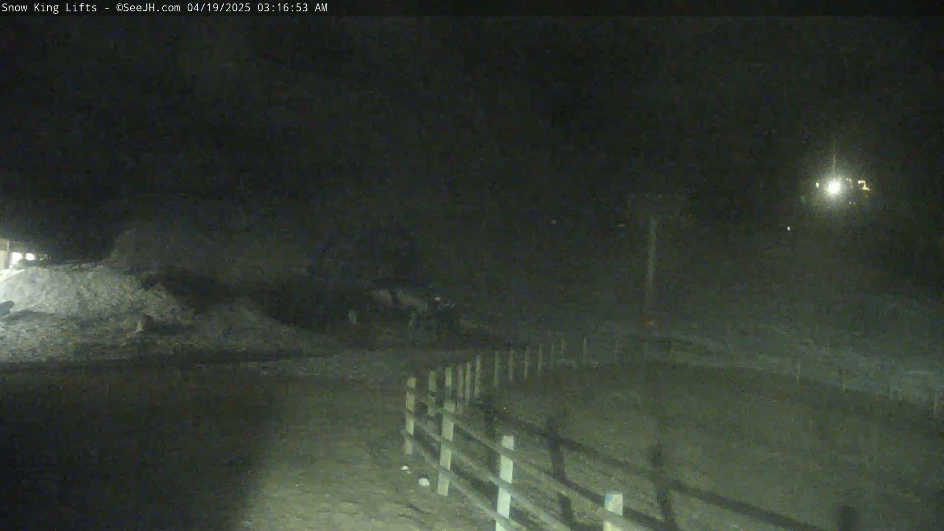 A dark, nighttime scene shows a snow-covered area with a wooden fence and a distant light source.