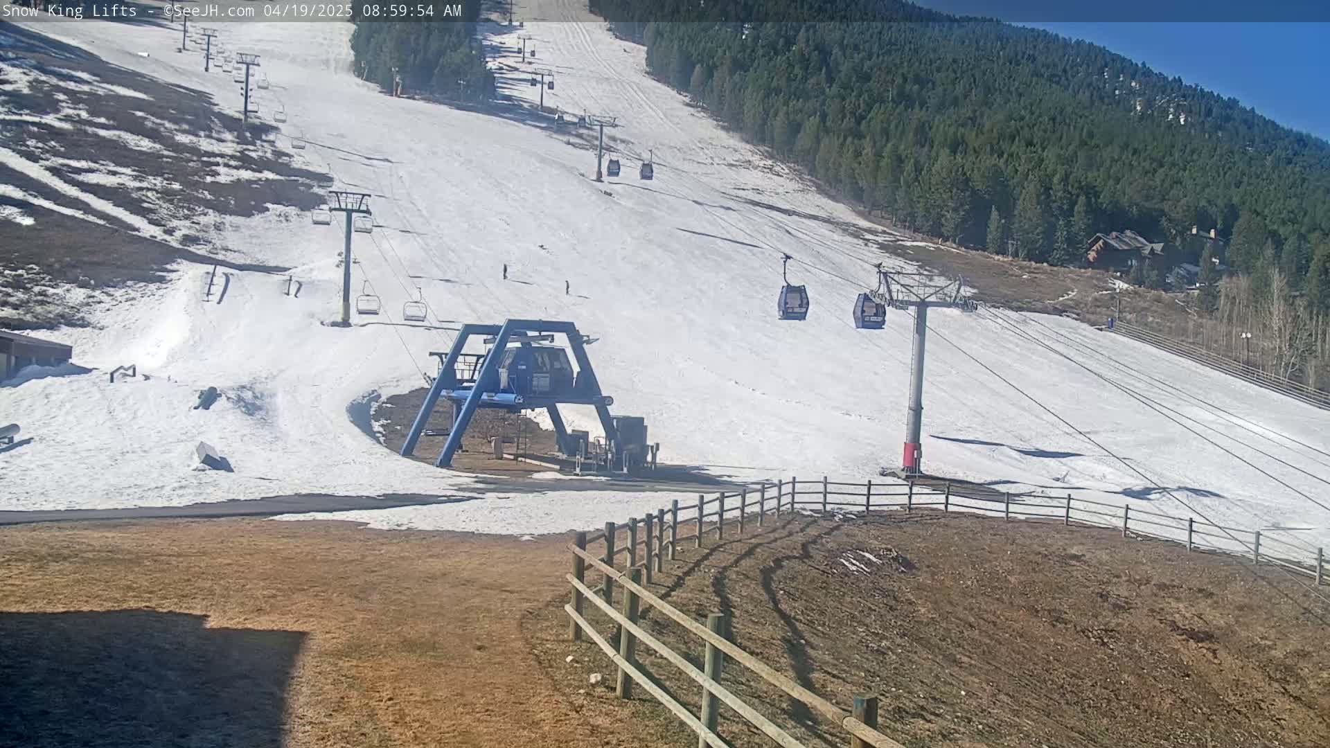 A ski lift base station and several chairlifts ascending a snow-covered ski slope on a sunny day, with a forested mountain in the background.