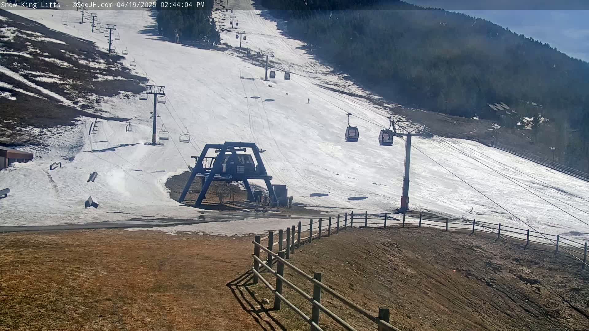 A ski lift and slope with patchy snow cover on a sunny day, bordered by a wooden fence and a background of trees.