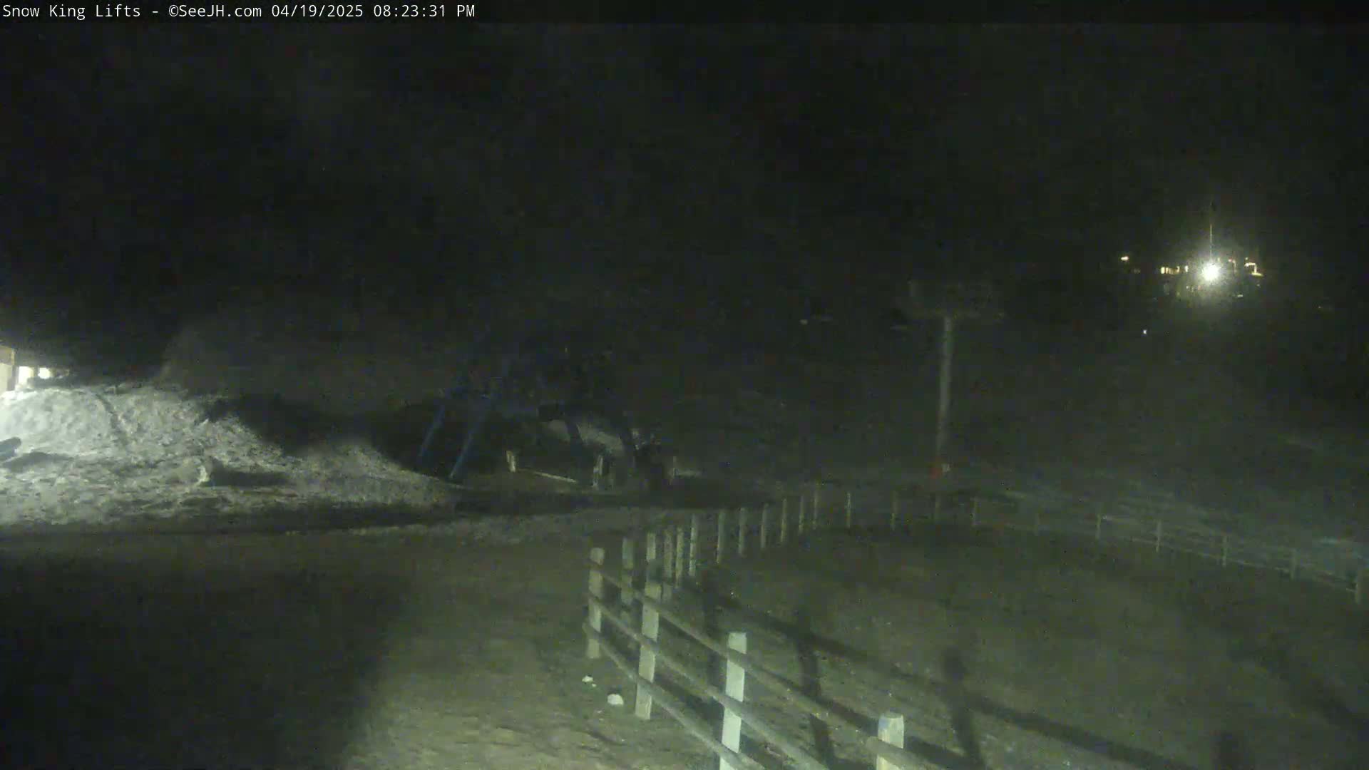 A nighttime, low-light view of a snow-covered area with a fence and a structure resembling a ski lift in the background.