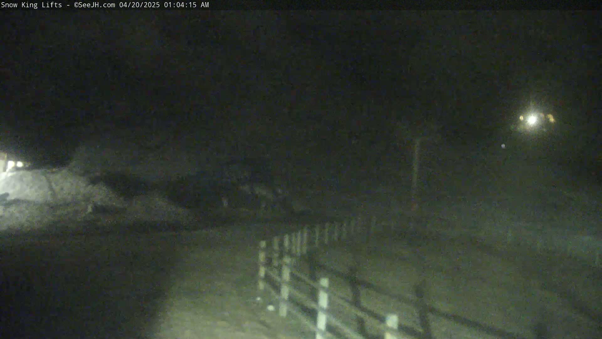 A nighttime, snow-covered scene shows a low-light view of a snow-covered area with a white fence, illuminated by a distant light source.