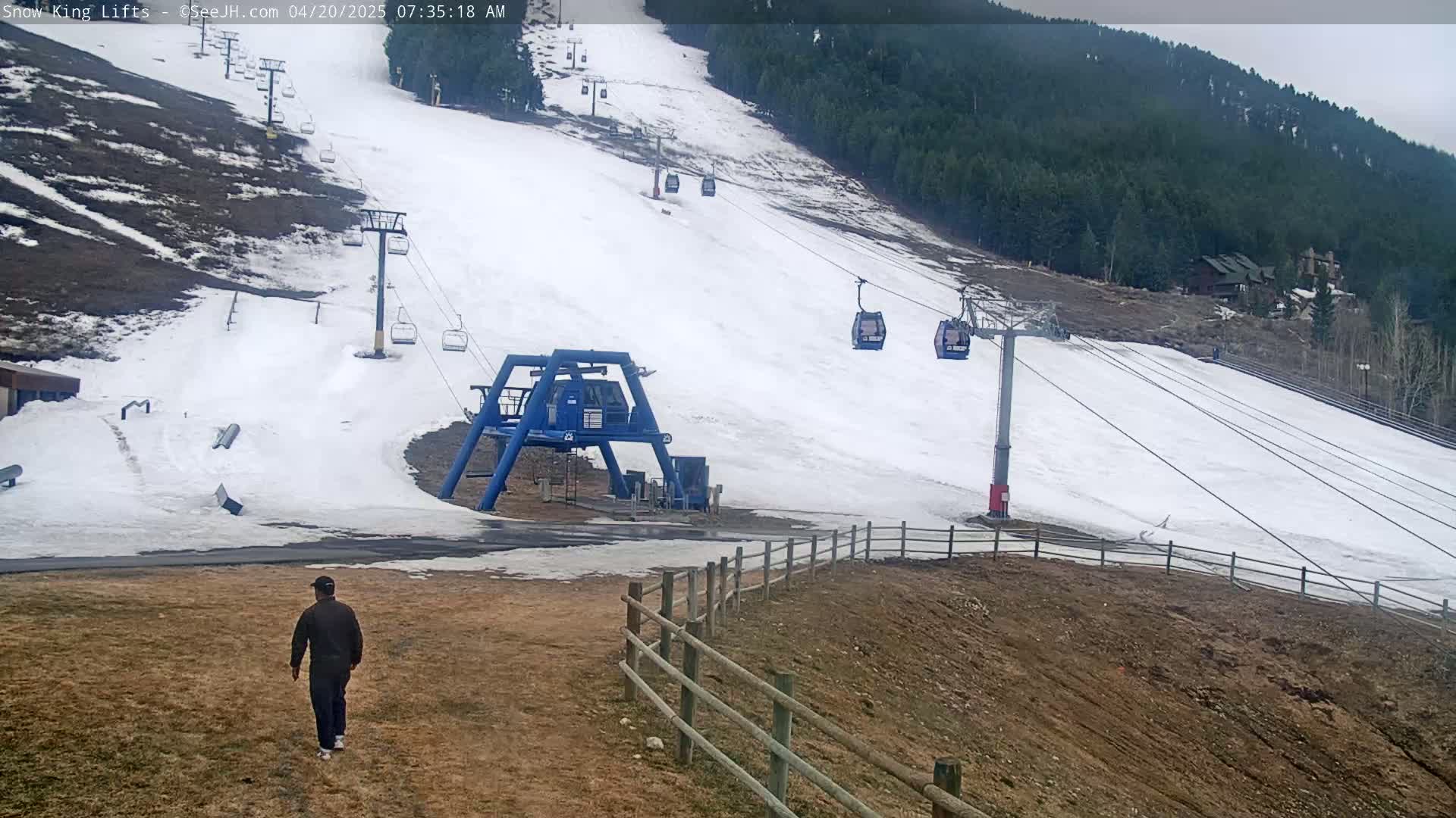 A person walks on a brown grassy area below a ski lift on a snowy hill under an overcast sky.