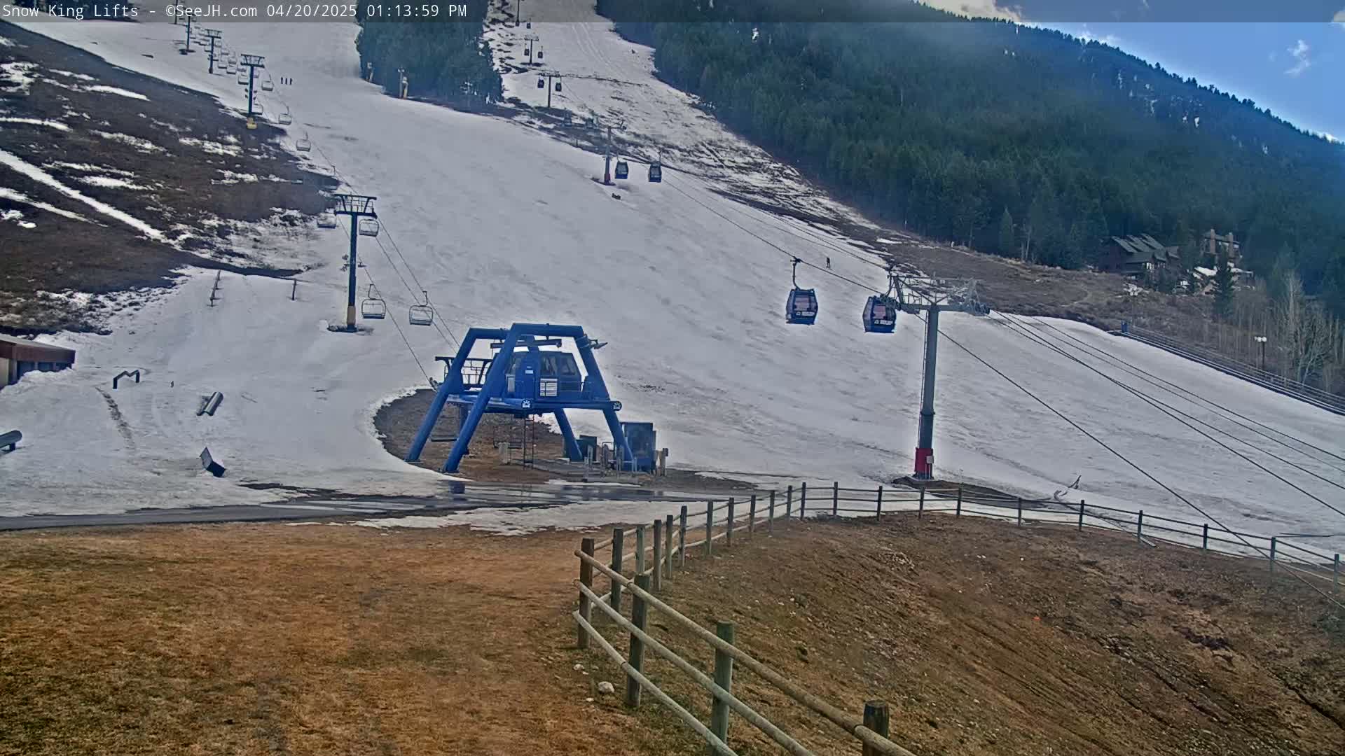 A ski lift with several empty chairlifts ascends a snow-covered slope on a partly cloudy day, with brown, barren areas visible at the bottom and sides.