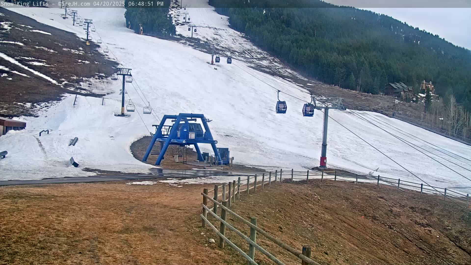 A ski lift with several empty chairlifts ascends a snow-covered slope on an overcast day, bordered by brown, barren ground and a distant evergreen forest.