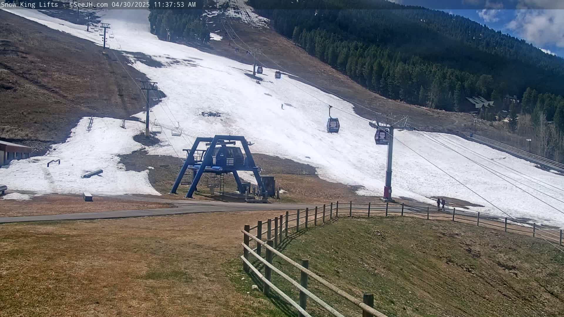A ski lift with several empty gondolas ascends a snow-covered hill on a partly sunny day, with brown grass visible at the base and a forested mountain in the background.