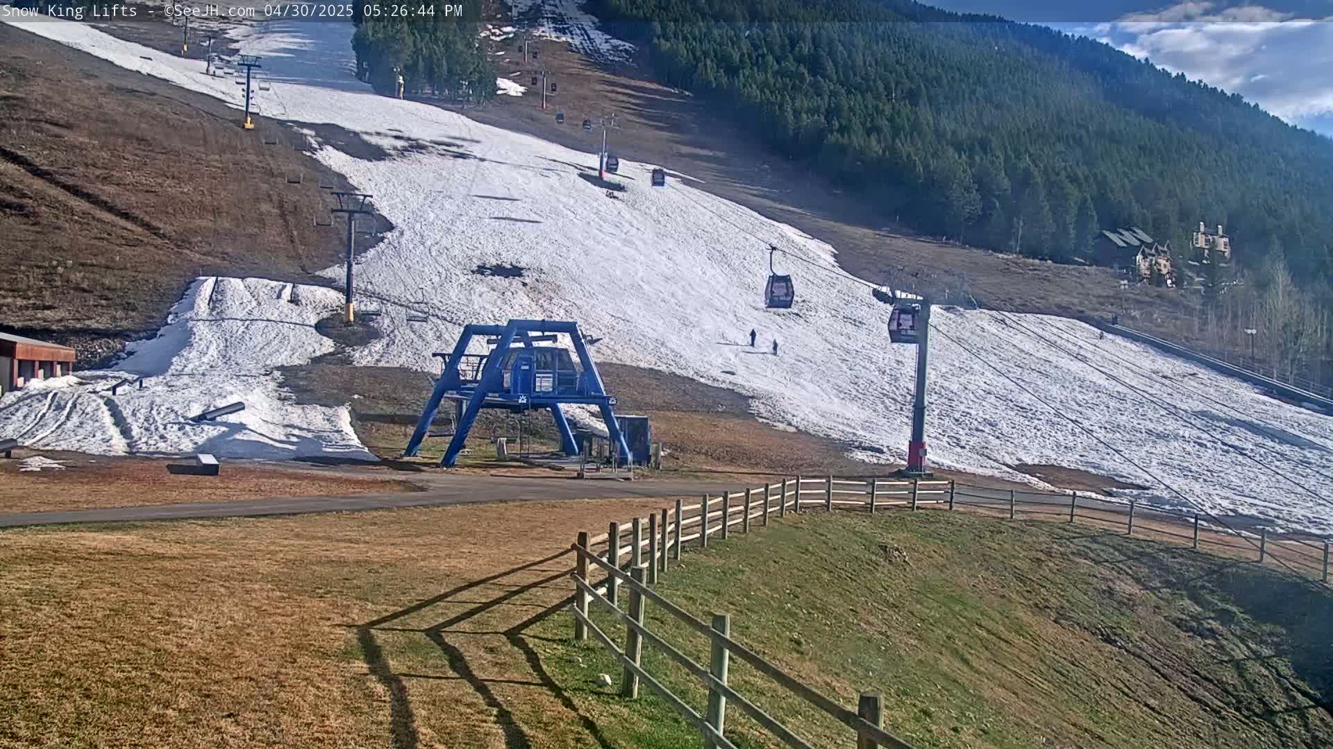 A ski slope with patchy snow, several chairlifts, and a few skiers under a sunny sky.