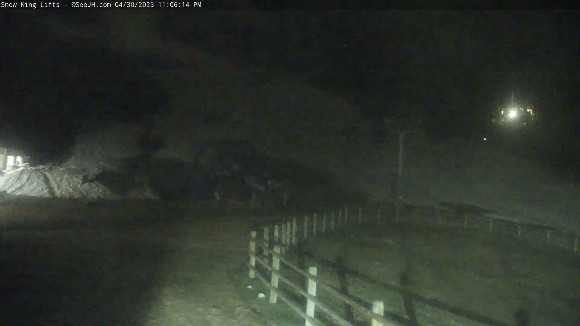A nighttime, snow-covered scene shows a pile of snow near a wooden fence under a dark sky.