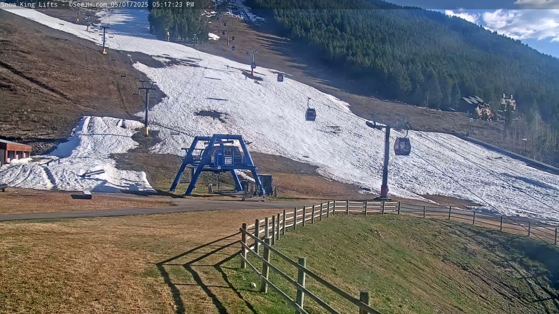 A ski lift with several empty chairs ascends a snow-covered slope on a sunny day, with a partially snow-covered grassy area in the foreground.