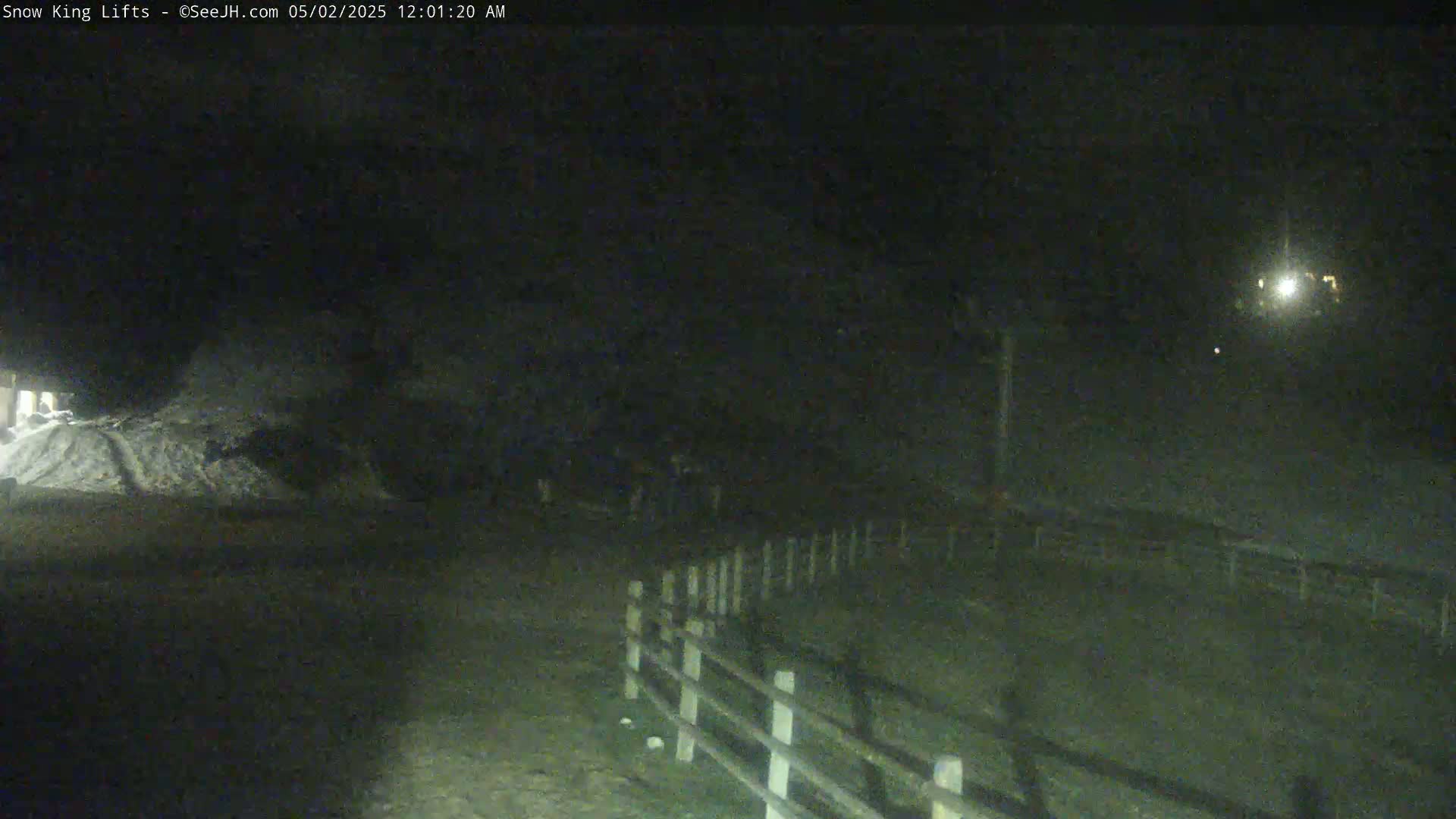 A nighttime, snow-covered scene shows a section of a wooden fence, a pile of snow, and indistinct figures in the distance under dark, possibly cloudy, skies.