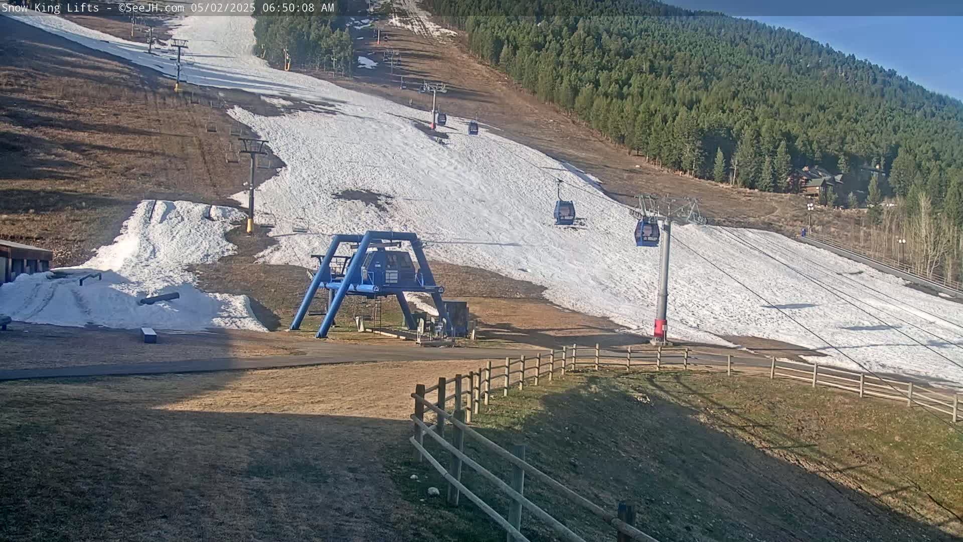 A ski lift with several empty gondolas ascends a snow-covered slope against a backdrop of a sunny, pine-covered mountain.