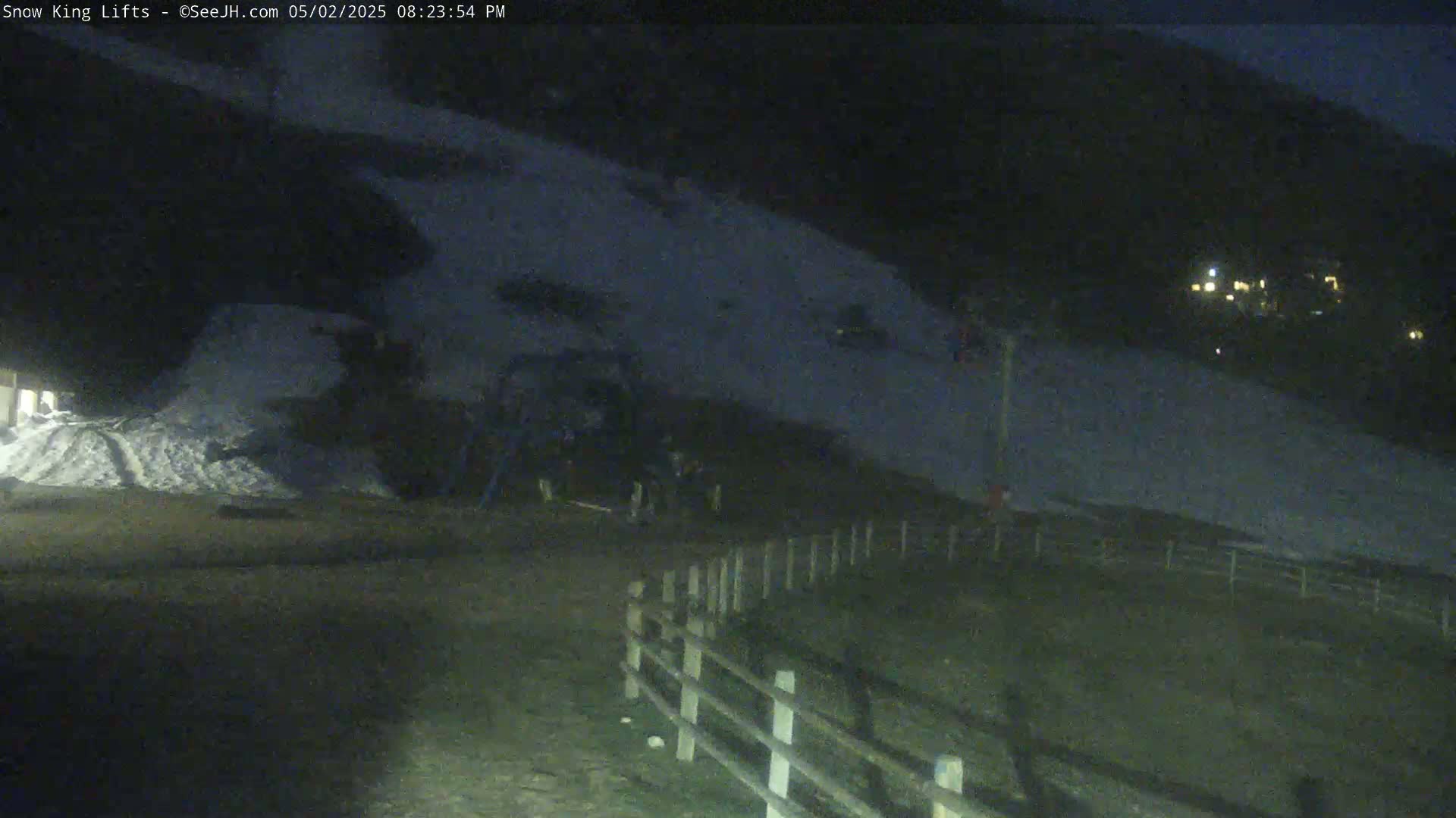 A nighttime, snow-covered slope is visible, with a wooden fence at the bottom and faint lights in the distance.