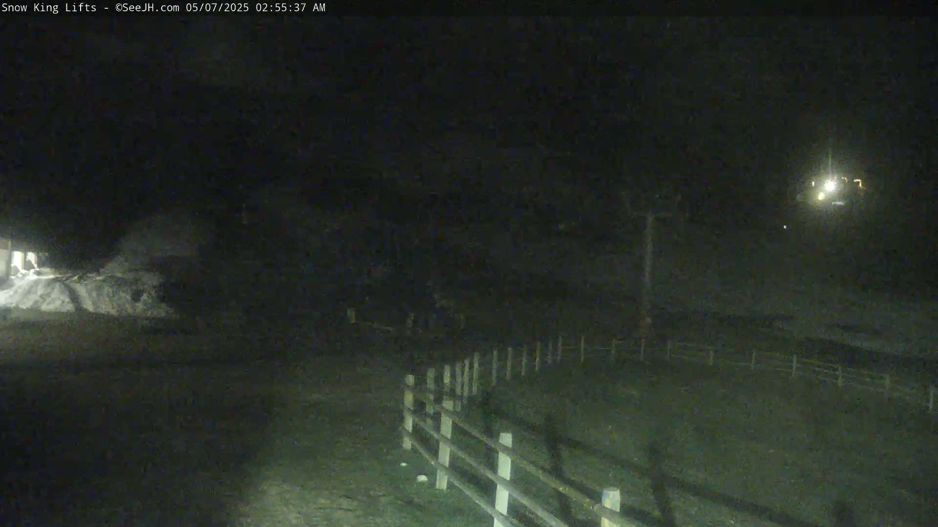 A dark, nighttime scene shows a snow pile, a wooden fence, and a distant light source.