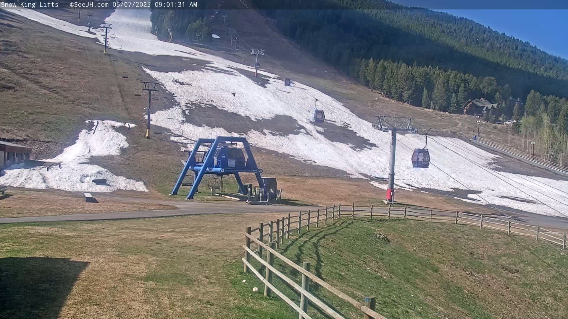 A ski lift with several empty gondola cars ascends a hill partially covered in snow under a sunny sky.