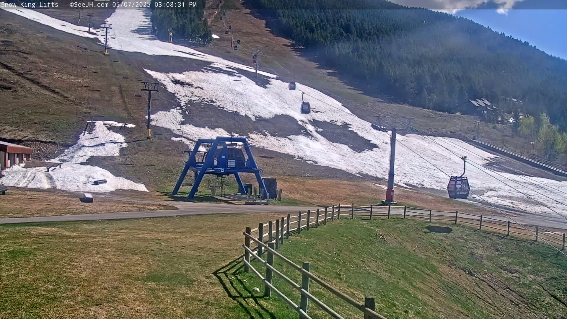 A ski lift with several passenger cars ascends a hill partially covered in snow under a partly sunny sky.