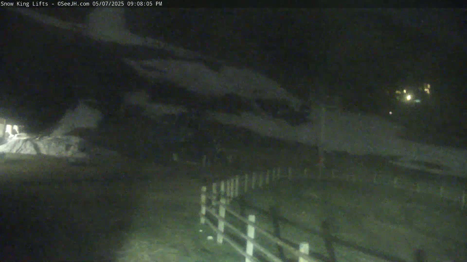 A nighttime view shows a snow-covered area with a fence and a dark building under a cloudy sky.