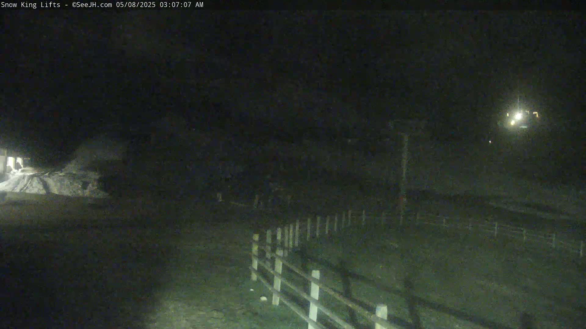 A nighttime, low-light view shows a snow-covered area near a fence, with a building visible in the background.