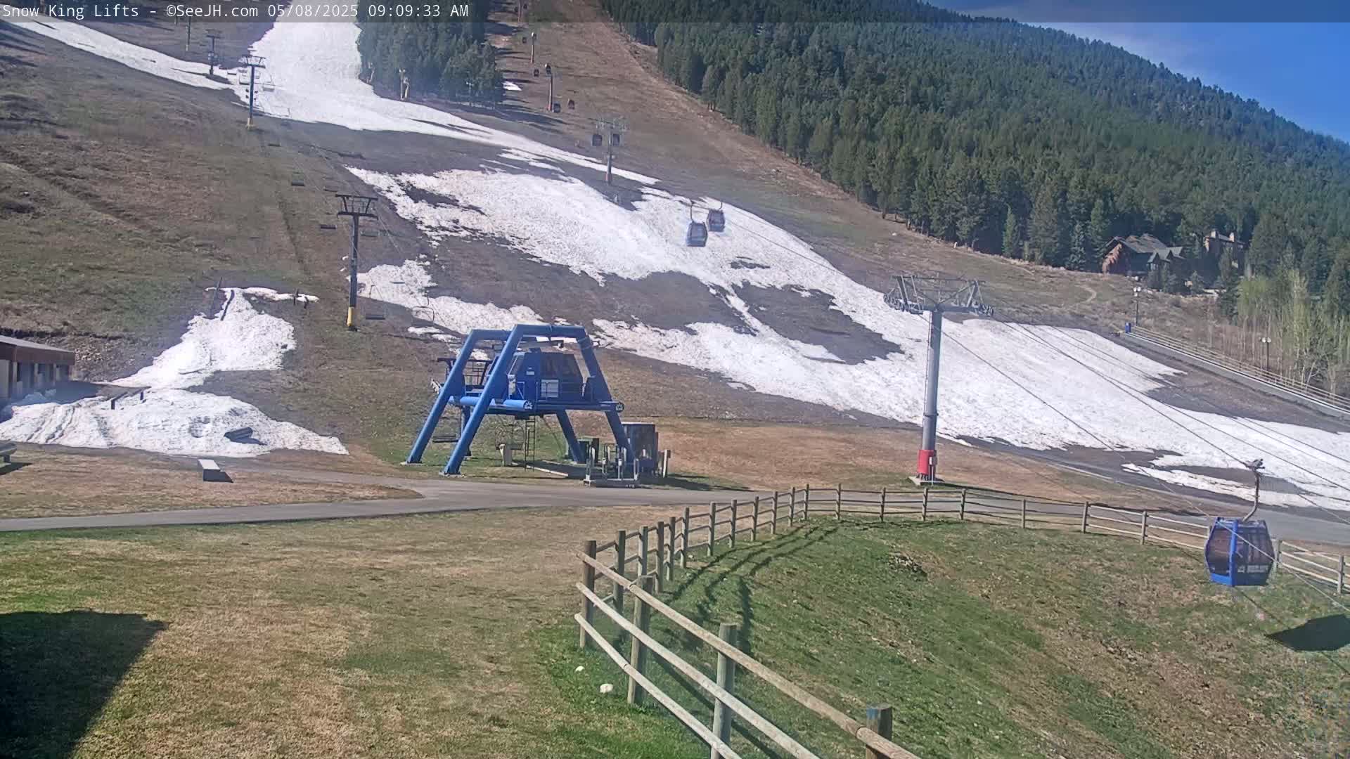 A mostly sunny view of a ski lift with patches of snow on a grassy hill, under a mostly clear sky.