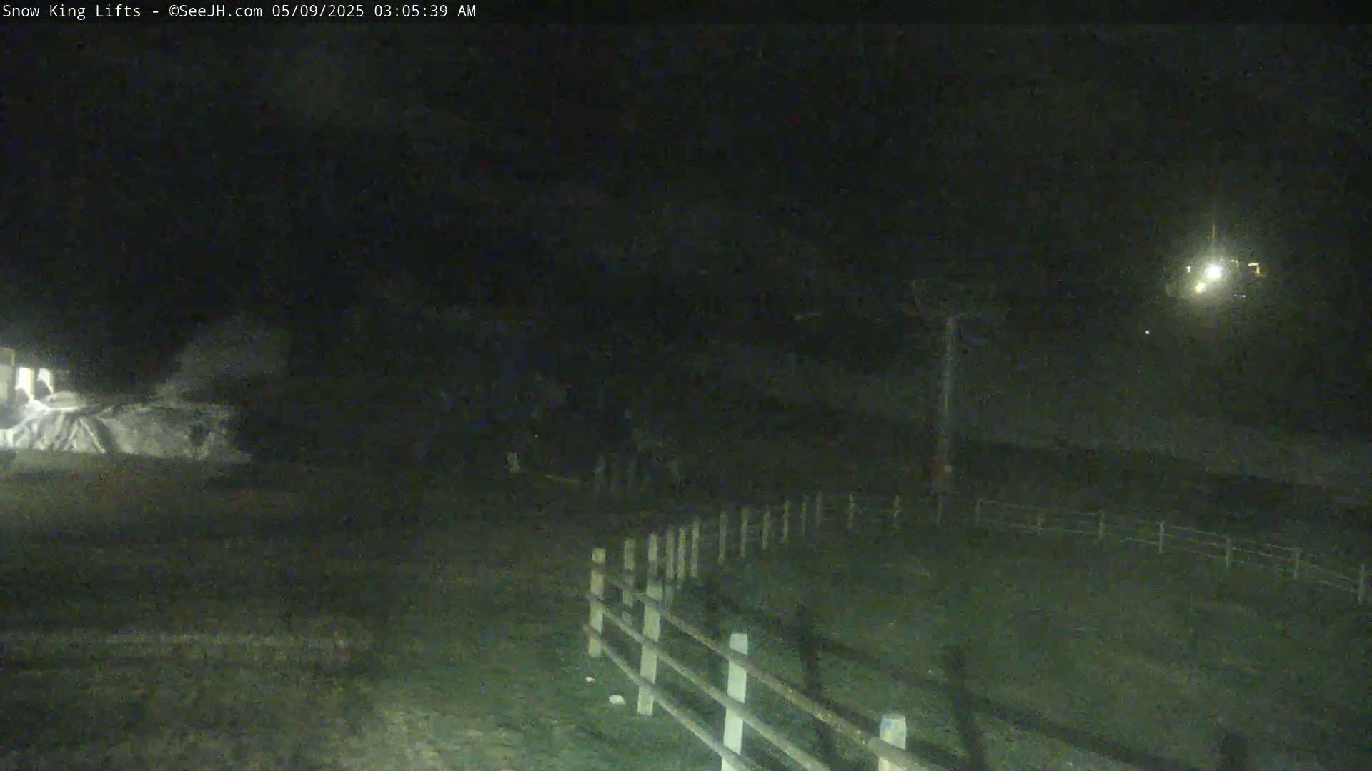 A nighttime, low-light view shows a snow-covered area near a wooden fence, with a dimly lit structure and a few indistinct figures in the distance.