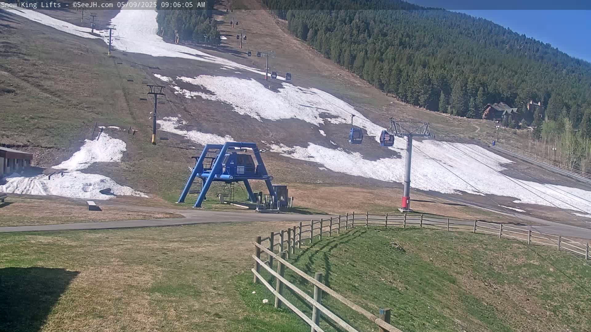 A ski lift and chairlifts are visible on a mostly bare ski slope with patches of snow under a sunny sky, surrounded by a grassy area and a wooden fence.