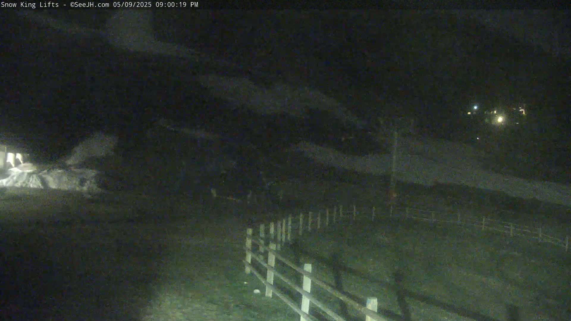 A nighttime, low-light view of a snow-covered area, possibly a ski resort, with a wooden fence and some dimly lit buildings in the distance.