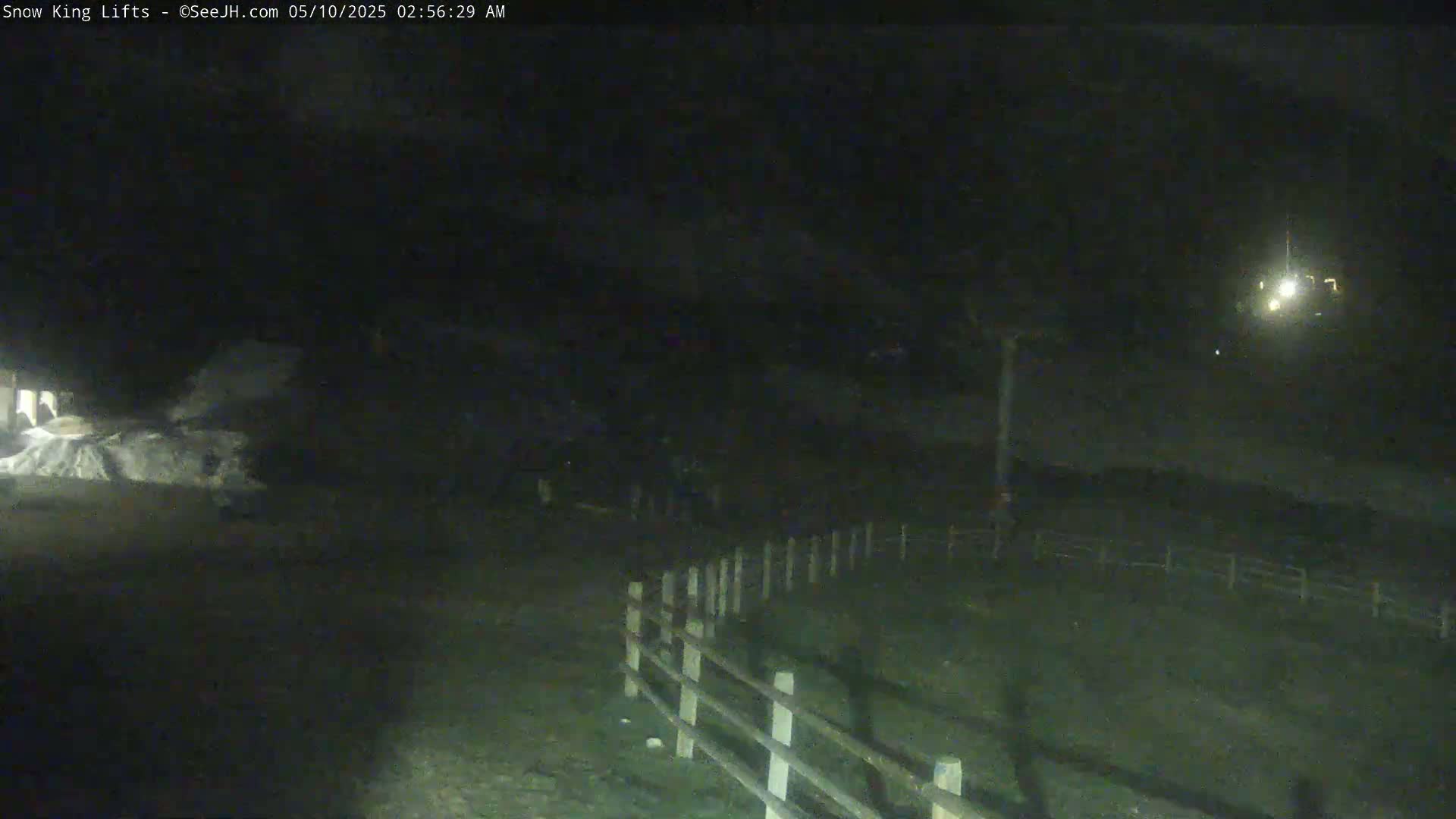 A nighttime, outdoor scene shows a dimly lit area with a wooden fence and a pile of what appears to be snow or ice in the distance.