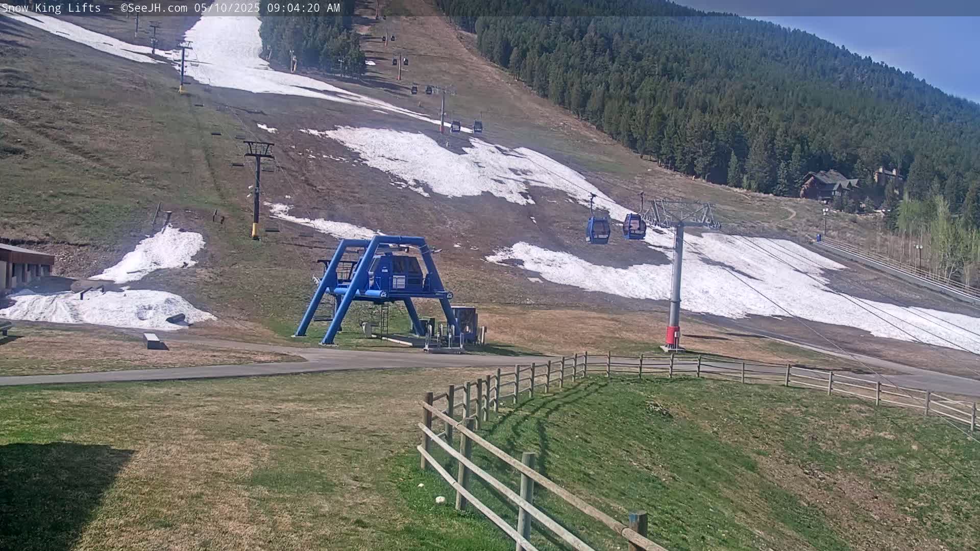 A mostly sunny view of a ski lift and mountainside with patches of remaining snow, grassy areas, and a wooden fence.