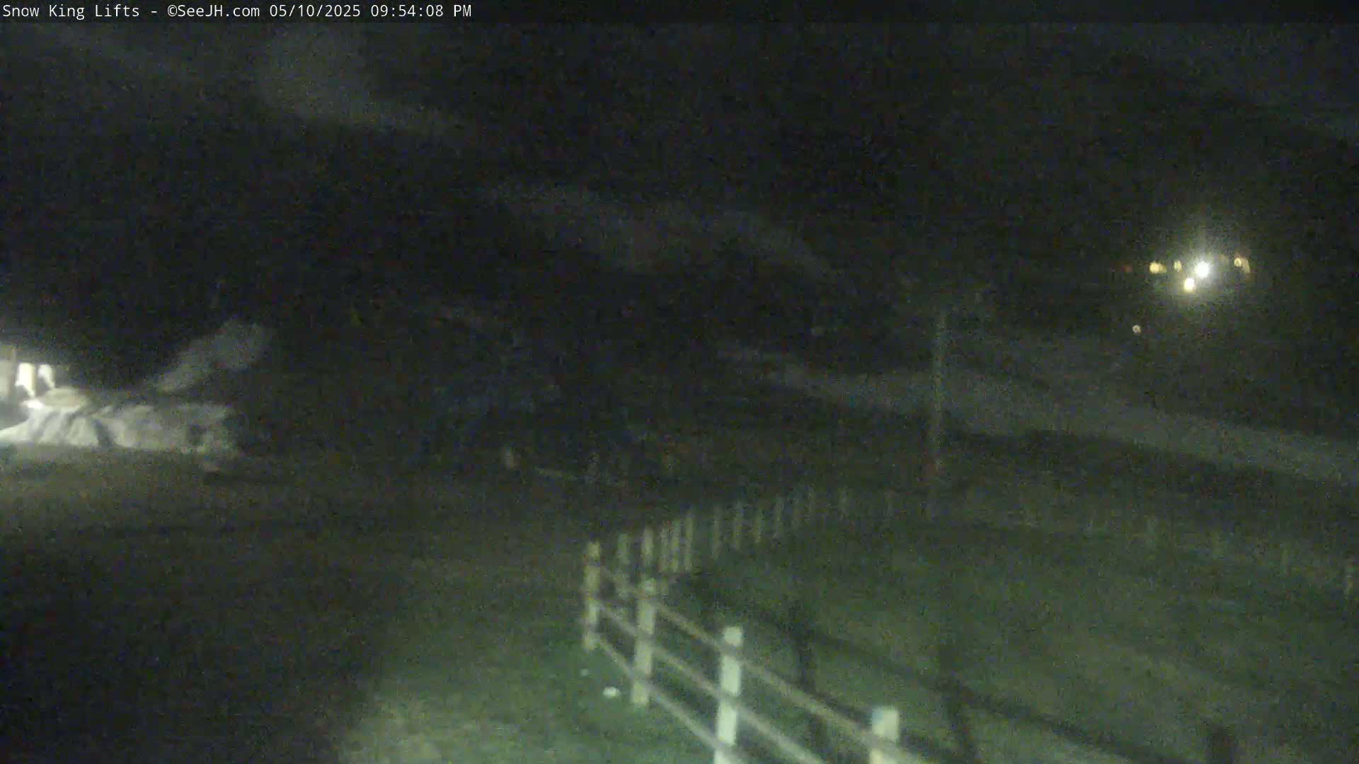 A nighttime, low-light view shows a snow-covered area near a fence, with some lights visible in the distance.